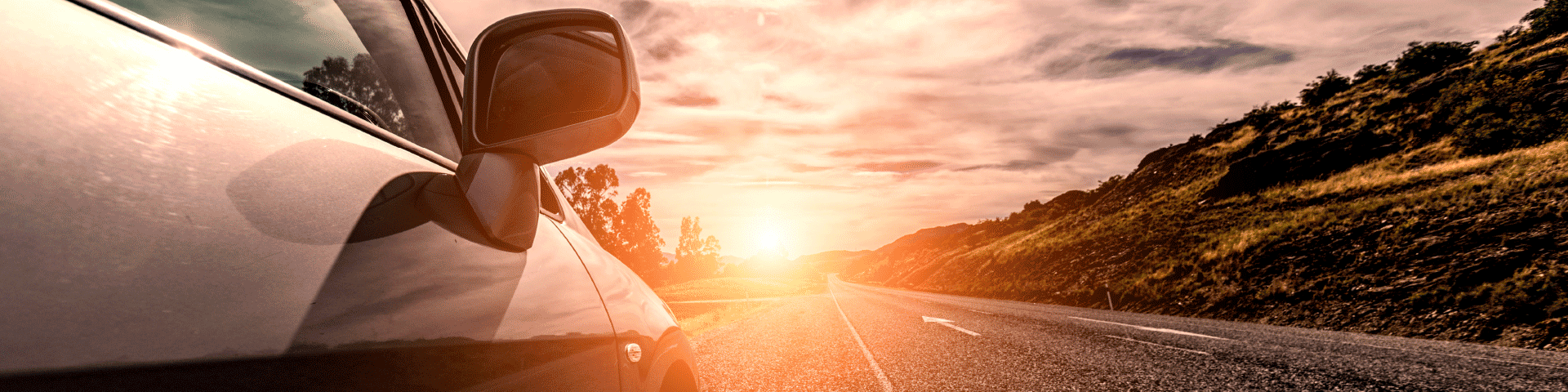 Image of a car driving down a highway with mountains on the right side and the sun setting.